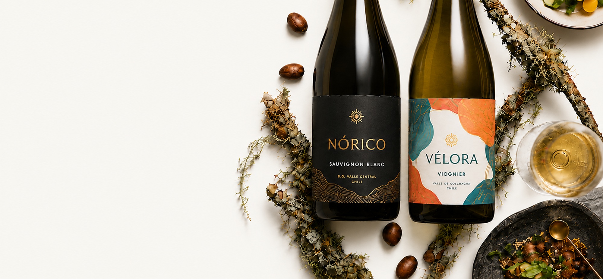 Botellas de vino Nórico Sauvignon Blanc y Vélora Viognier sobre una mesa blanca, acompañadas de una copa de vino y preparaciones gastronómicas como propuesta de maridaje.